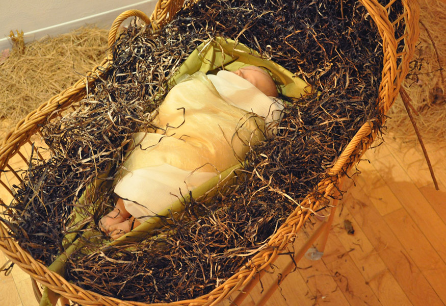 COCOONS Embryonic humans are developing as they dangle from the ceiling, coming to life in the manner of insects. Dolls, fabric, branches, bassinet, seagrass, pine needles